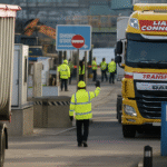 A yellow Liam Connolly Transport truck approaches a port checkpoint where a worker in a high-visibility jacket signals traffic amid industrial surroundings.